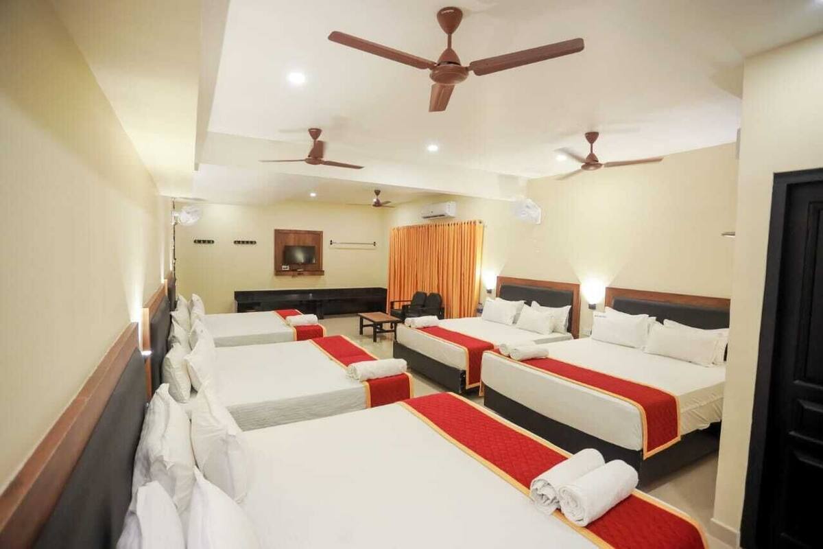 A hotel room with four single beds, each with a red bedspread and white pillows. There are ceiling fans, a television, and a desk with a chair.