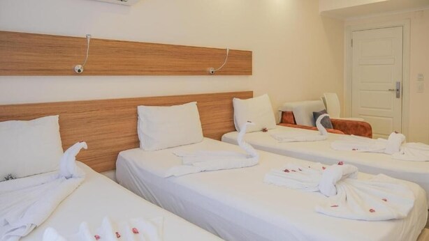Ein Hotelzimmer mit drei Betten, jeweils mit weißen Laken und Handtüchern. Über den Betten ist ein hölzerner Kopfteil zu sehen, und an der Wand ist eine Klimaanlage angebracht.