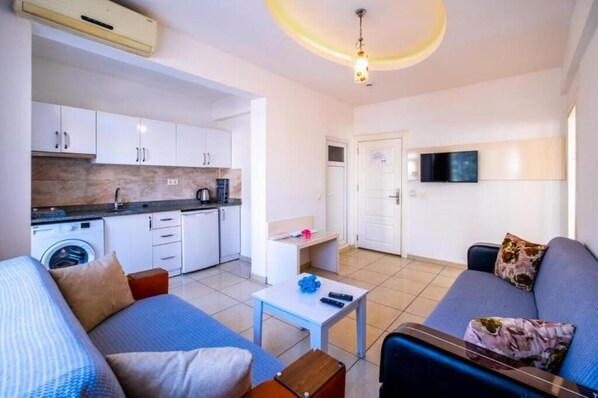 Un espacio habitable compacto con kitchenette, un sofá, una mesita y un televisor montado en la pared.
