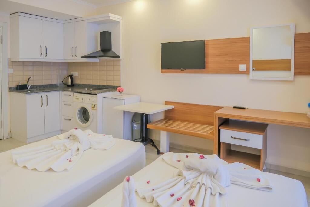 A hotel room with a kitchenette, a washing machine, a bed with a wooden headboard, a bedside table, and a mirror.