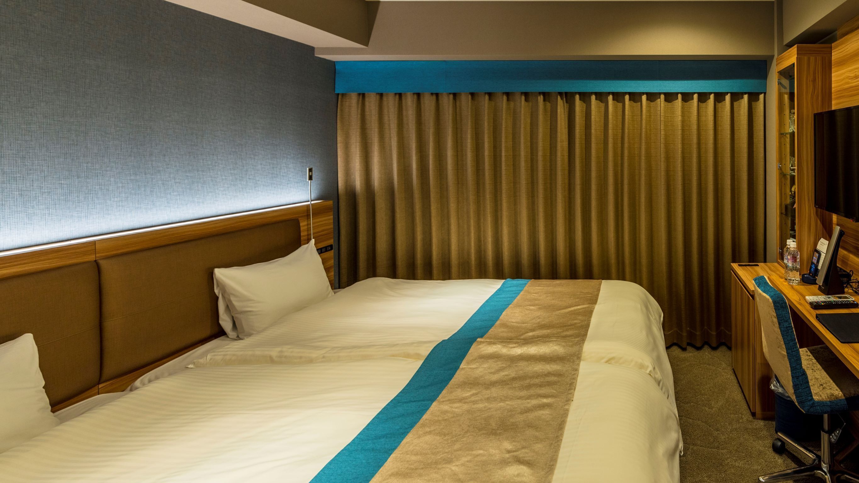 A hotel room with a bed, a desk with a computer, a chair, and a window with curtains.