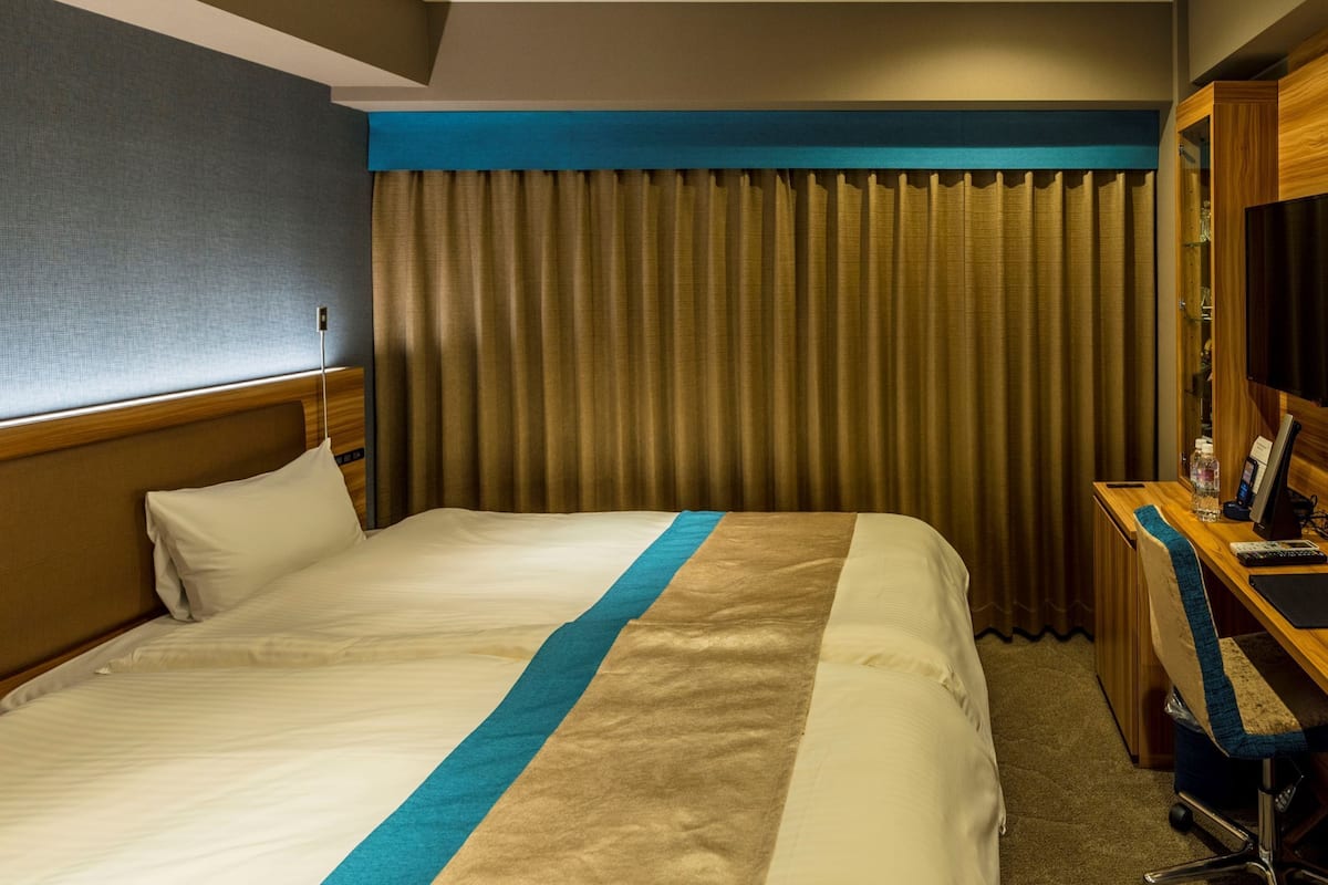 A hotel room with a bed, a desk with a computer, a chair, and a window with curtains.