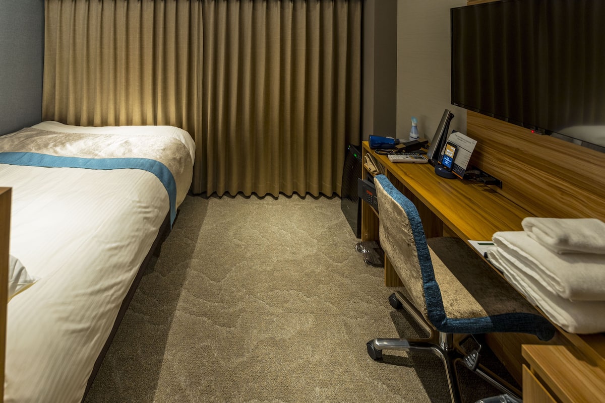 A hotel room with a bed, desk, chair, and a large window with curtains.