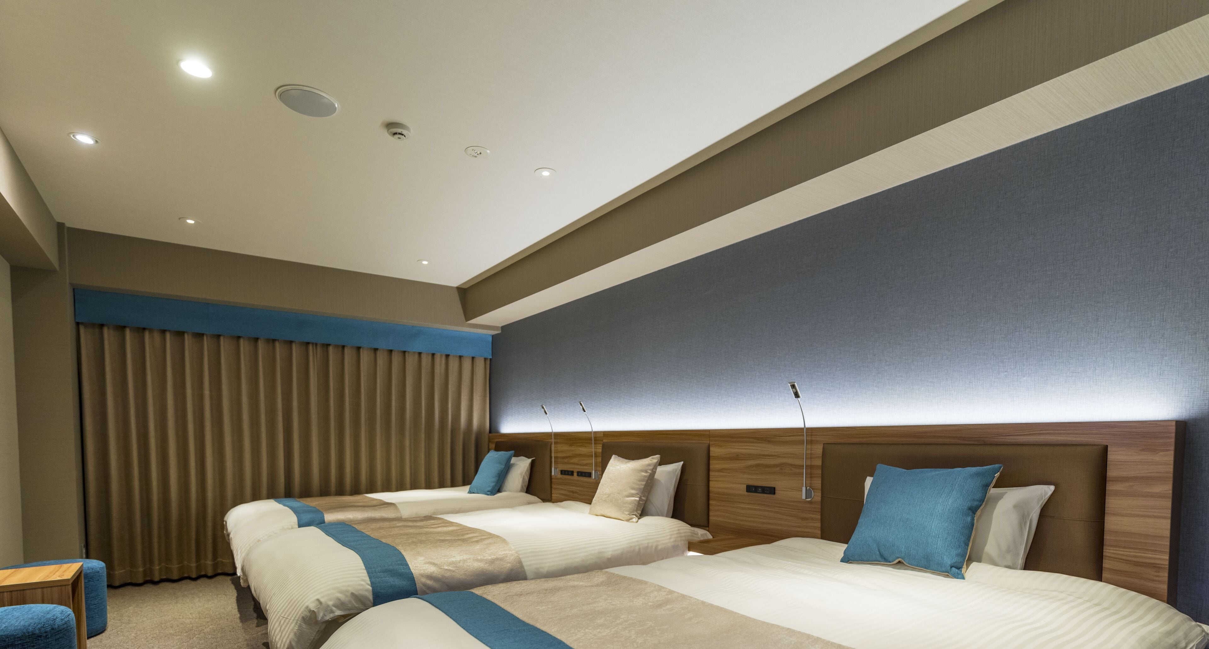 Habitación de hotel con dos camas, un ventanal con cortinas, cabecero de madera e iluminación empotrada en el techo.