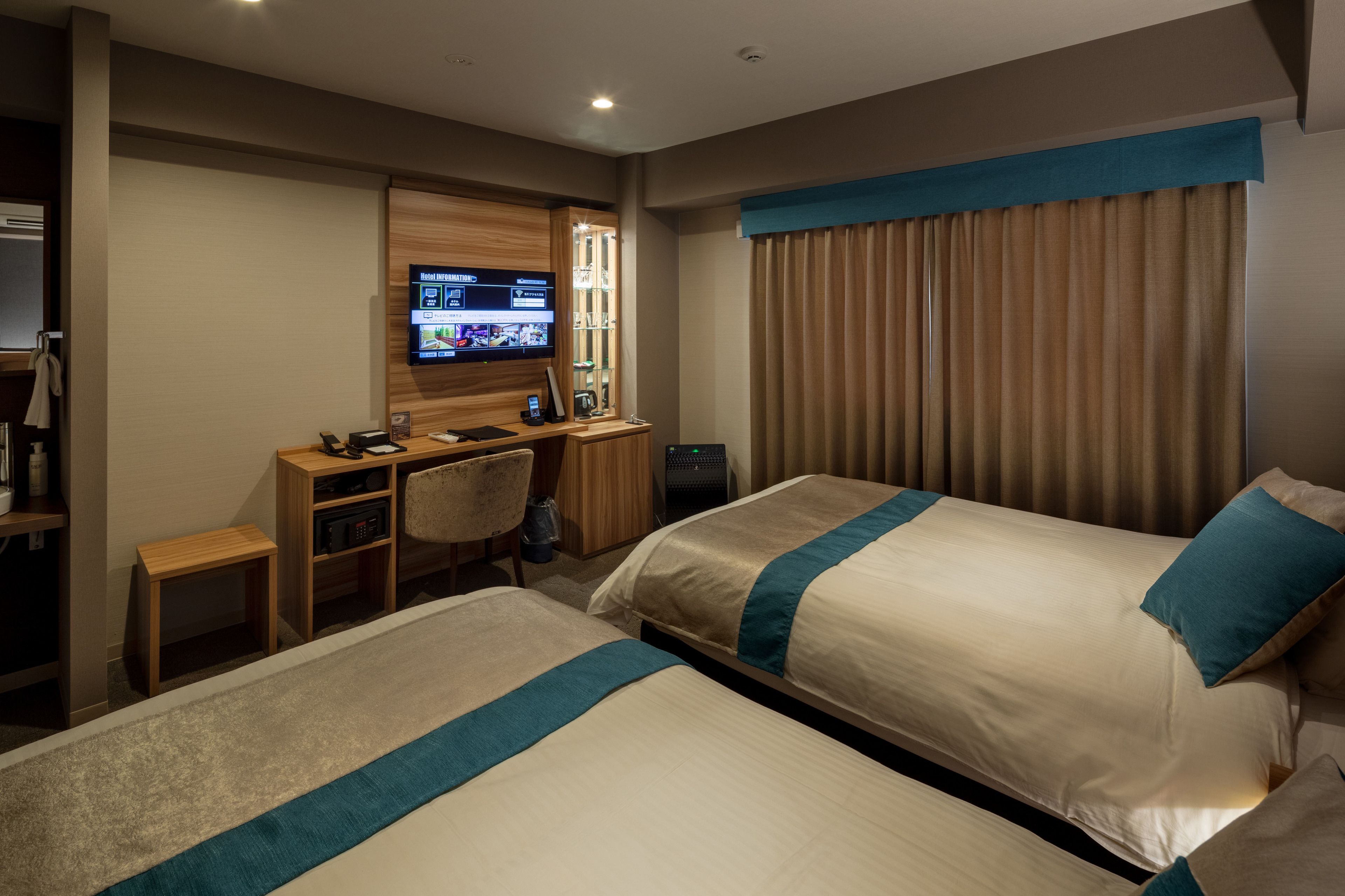 Ein Hotelzimmer mit zwei Betten, einem Schreibtisch mit Computer, einem Fernseher und einem Fenster mit Vorhängen.