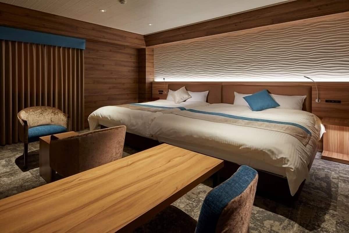 A hotel room with a large bed, a wooden desk, two chairs, and a window with vertical blinds.
