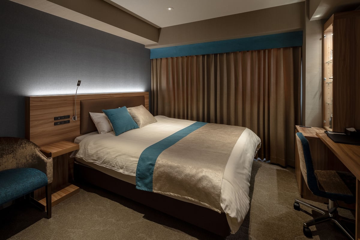 A hotel room with a bed, a desk, a chair, a nightstand, and a wardrobe.
