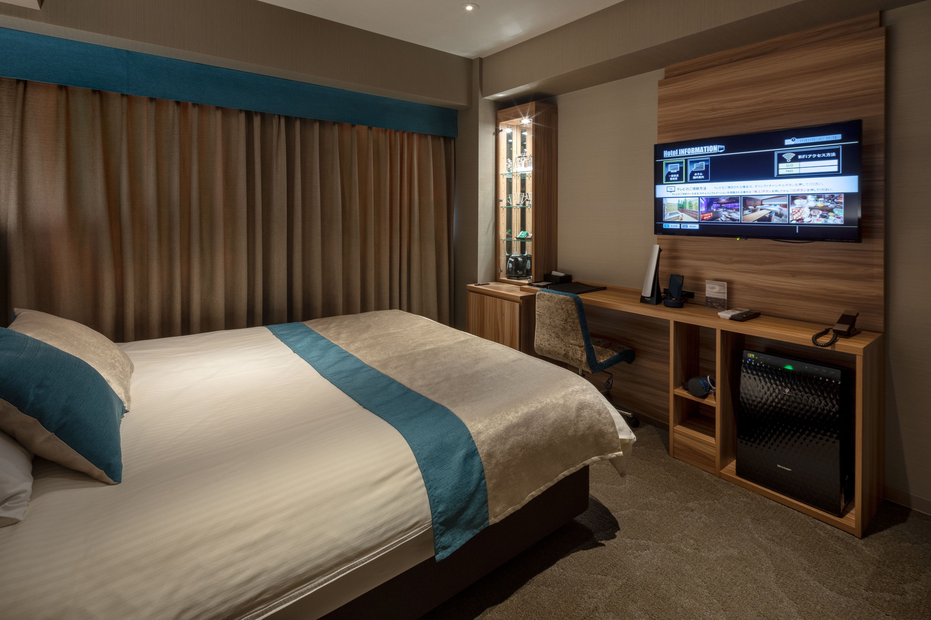 A hotel room with a bed, a desk with a computer, a TV on the wall, and a minibar.