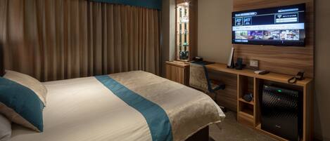 A hotel room with a bed, a desk with a computer, a TV on the wall, and a minibar.