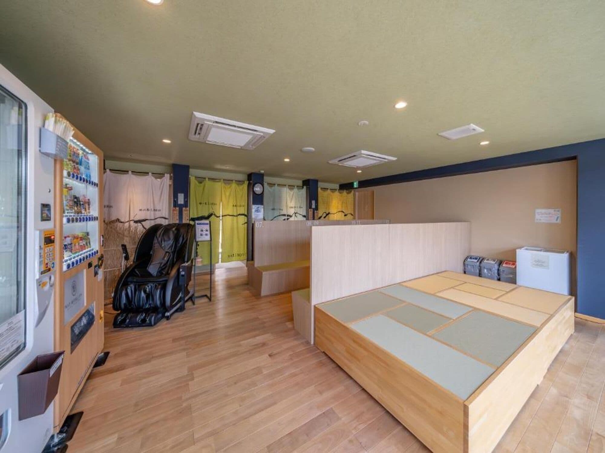 A room with a wooden floor, a low wooden bed, a black chair, a vending machine, and a small refrigerator.