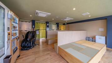 A room with a wooden floor, a low wooden bed, a black chair, a vending machine, and a small refrigerator.