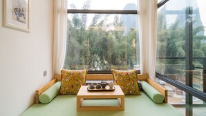 A room with a green bed, floral pillows, a wooden table with tea sets, and a view of trees through large windows. - CTN LiRiver Boutique Hotel (Guilin)