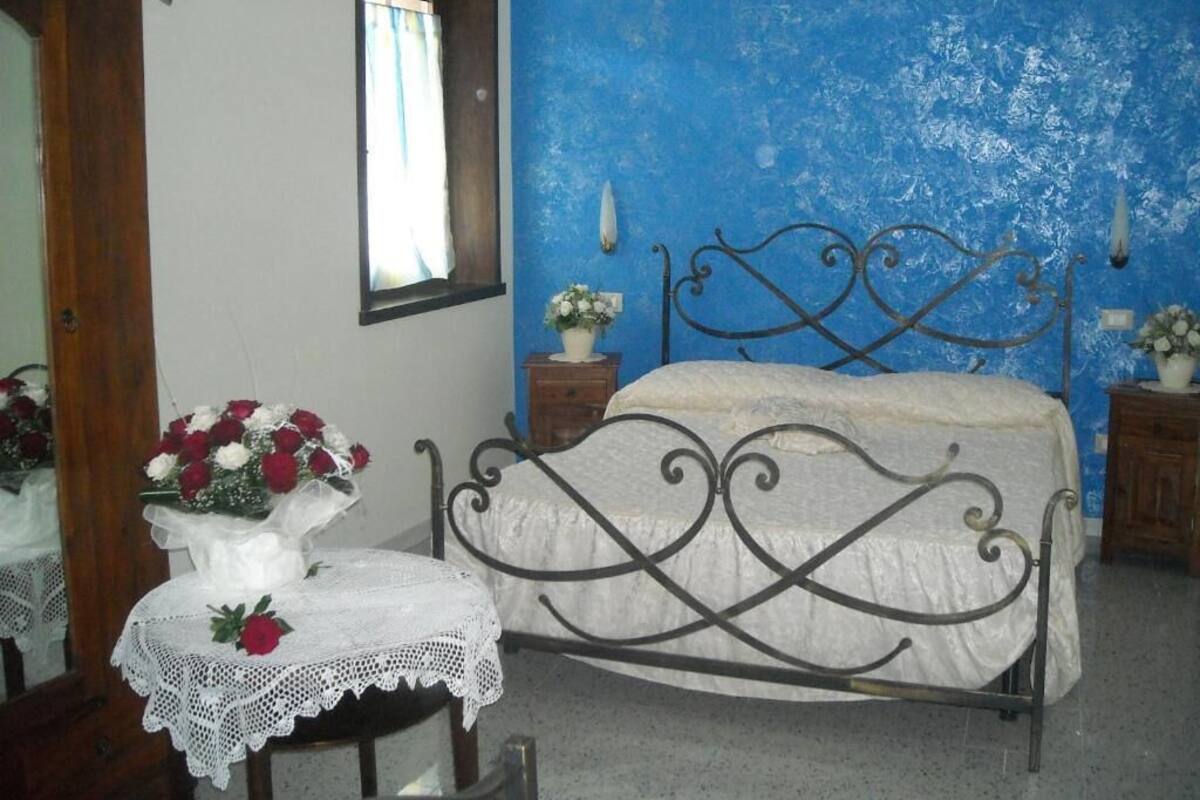 A bedroom with a blue wall, a metal bed frame, a wooden nightstand, and a small table with a lace tablecloth and a vase of flowers.