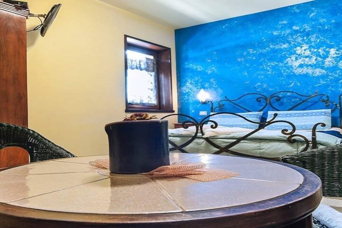 A room with a bed, a round table, wicker furniture, a mirror, and a blue wall.