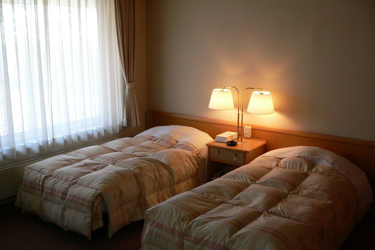 Una habitación de hotel con dos camas individuales, una mesita de noche con lámpara y una ventana con cortinas translúcidas.