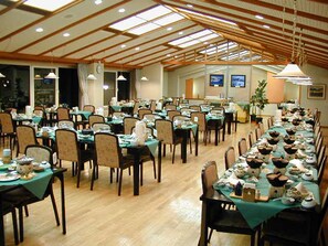 A large dining hall with long tables set for a meal, wooden beams on the ceiling, and a view of the outdoors through large windows. - Kusatsu Green Park Palace (Kusatsu)