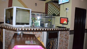 A kitchen area with a refrigerator, a microwave, a glass display case with fruits, a coffee machine, and a television mounted on the wall. - Sir Kanja's Hotel (Nairobi)