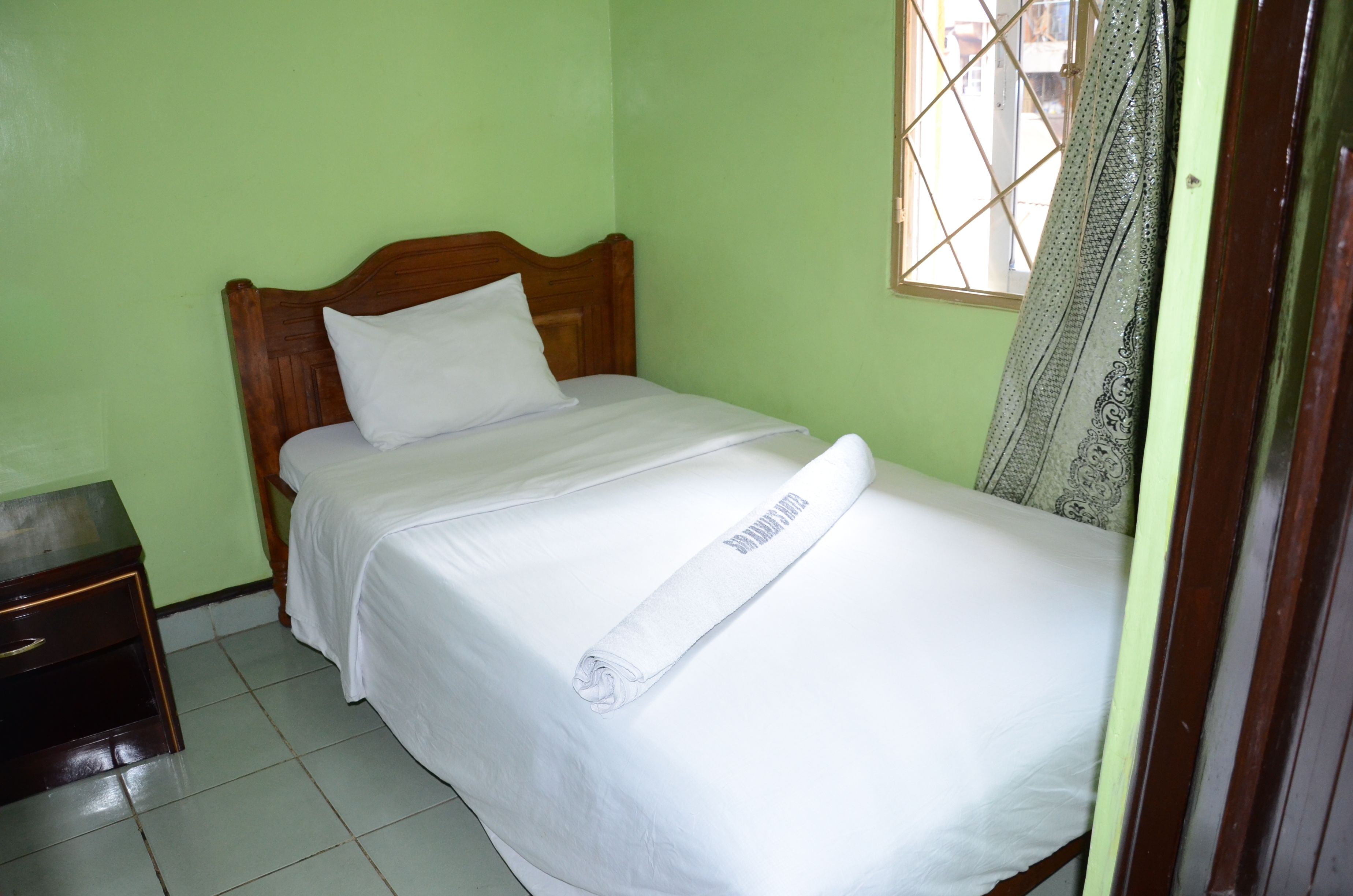 A single bed with a wooden headboard, a white pillow, and a neatly folded white towel on the bed. There is a small wooden nightstand to the left of the bed.
