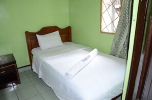 A single bed with a wooden headboard, a white pillow, and a neatly folded white towel on the bed. There is a small wooden nightstand to the left of the bed.