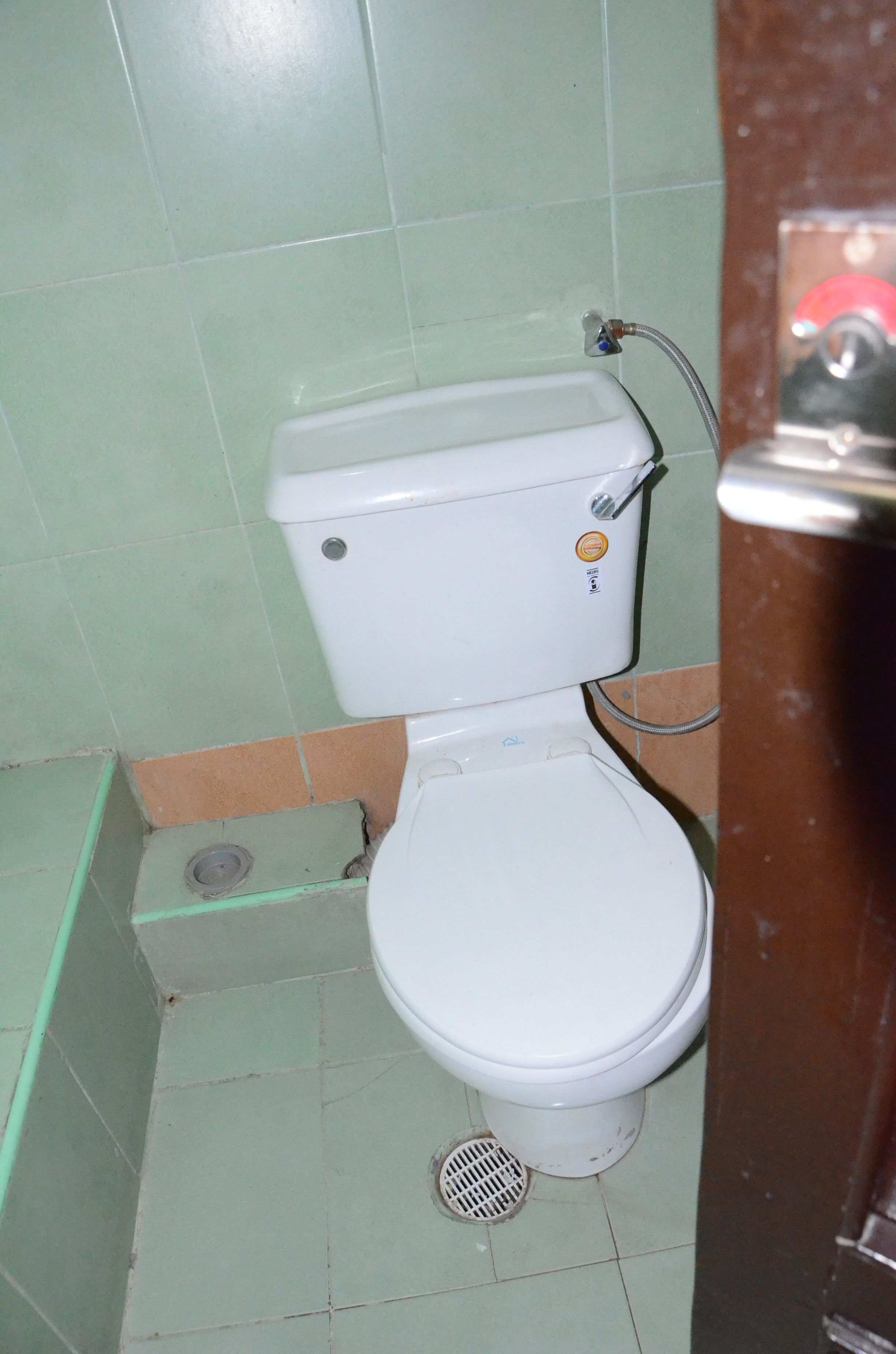 A small, basic bathroom with a toilet, a visible drain, and a tiled floor.