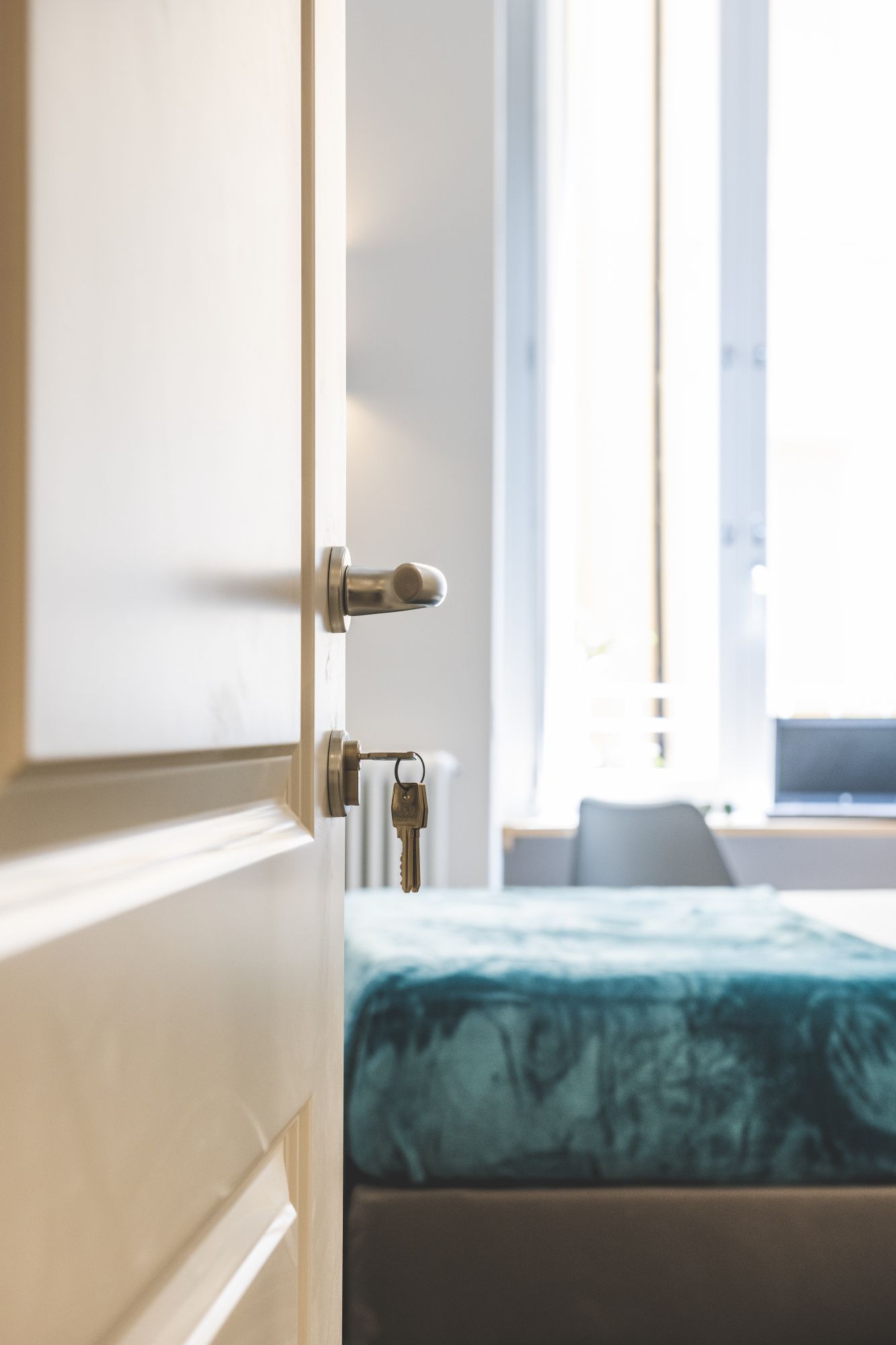 A hotel room with a bed, a door with a key in the lock, and a window with curtains.