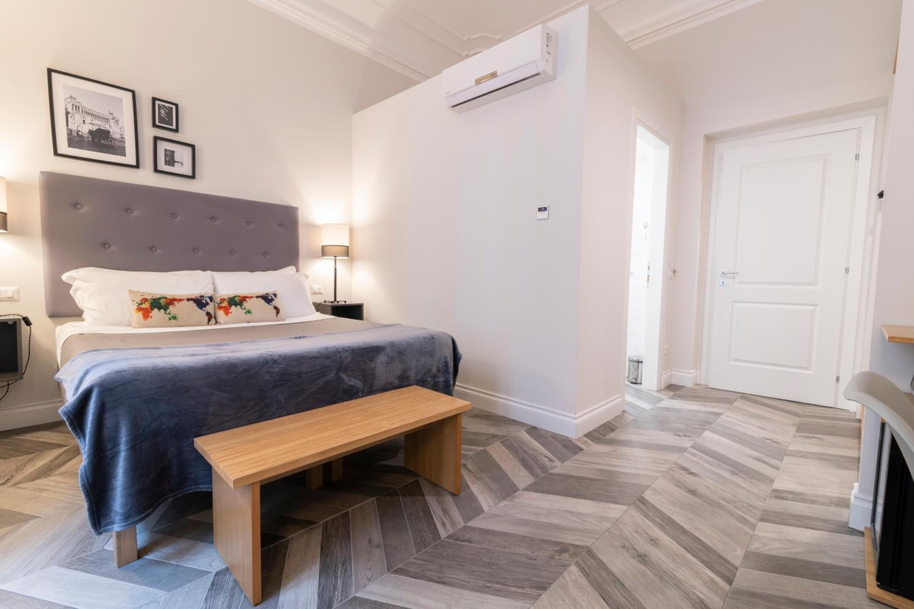 Une chambre à coucher comprenant un lit, des tables de chevet, un banc, un climatiseur fixé au mur et une porte.