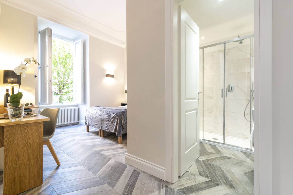 Hotellihuone, jossa on sänky, työpöytä lampun kanssa, ikkuna, josta on näkymä vehreään maisemaan, ja suihkutila.