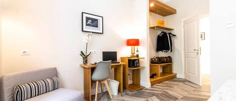 Habitación de hotel con sofá, escritorio con computadora, lámpara, estante con ropa y zapatos, y una puerta.