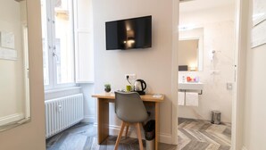 Quarto moderno com escrivaninha de madeira, uma cadeira, televisão montada na parede, radiador, janela, pia e lixeira.
