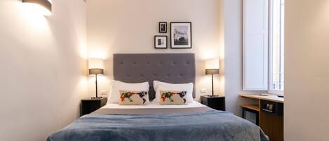 A neatly arranged bedroom with a large bed, two bedside tables with lamps, a window, and two framed pictures on the wall.