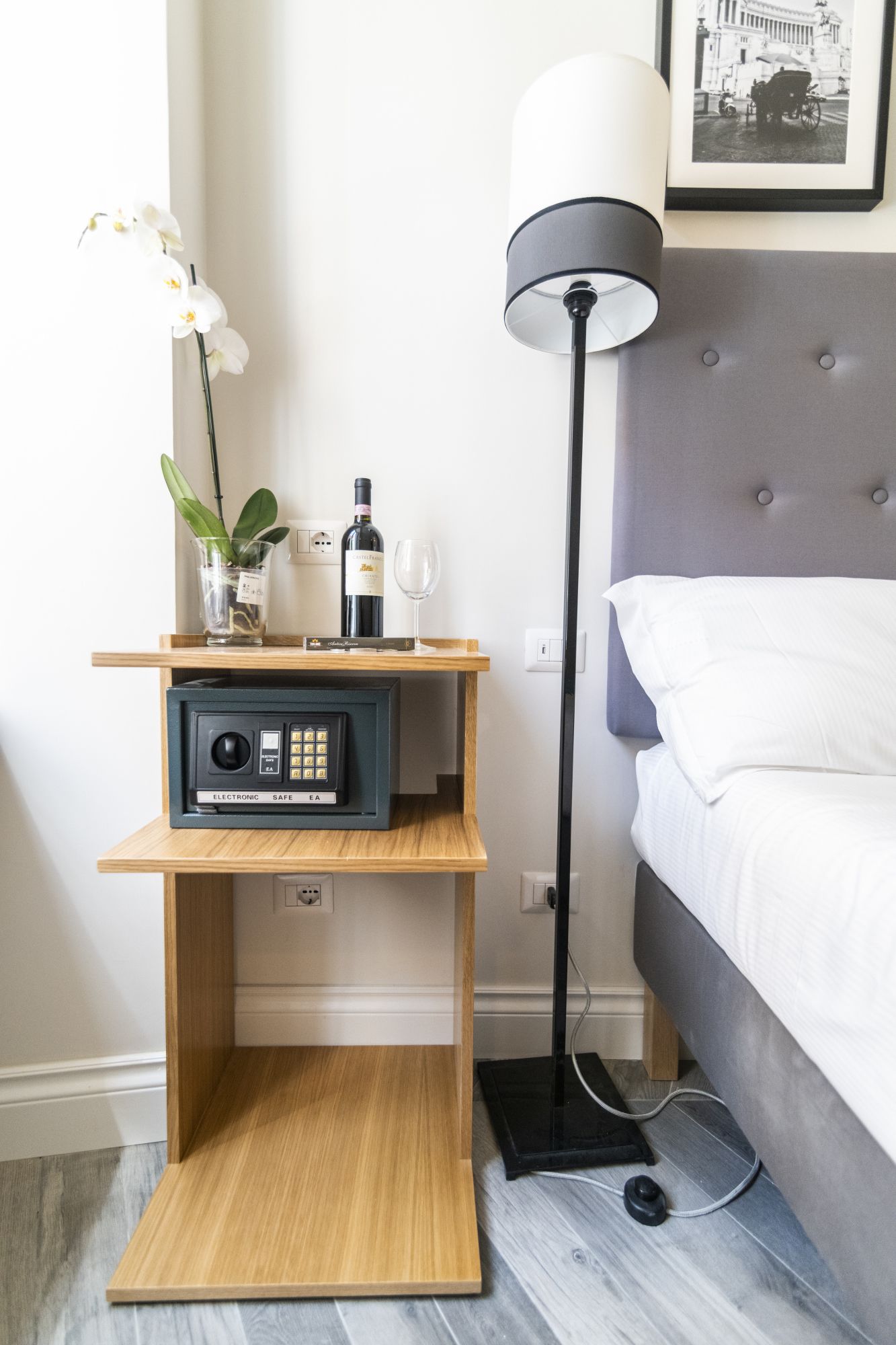 A neatly arranged hotel room with a bedside table holding a wine bottle, a glass, and a small plant. A floor lamp and a framed picture are also visible.