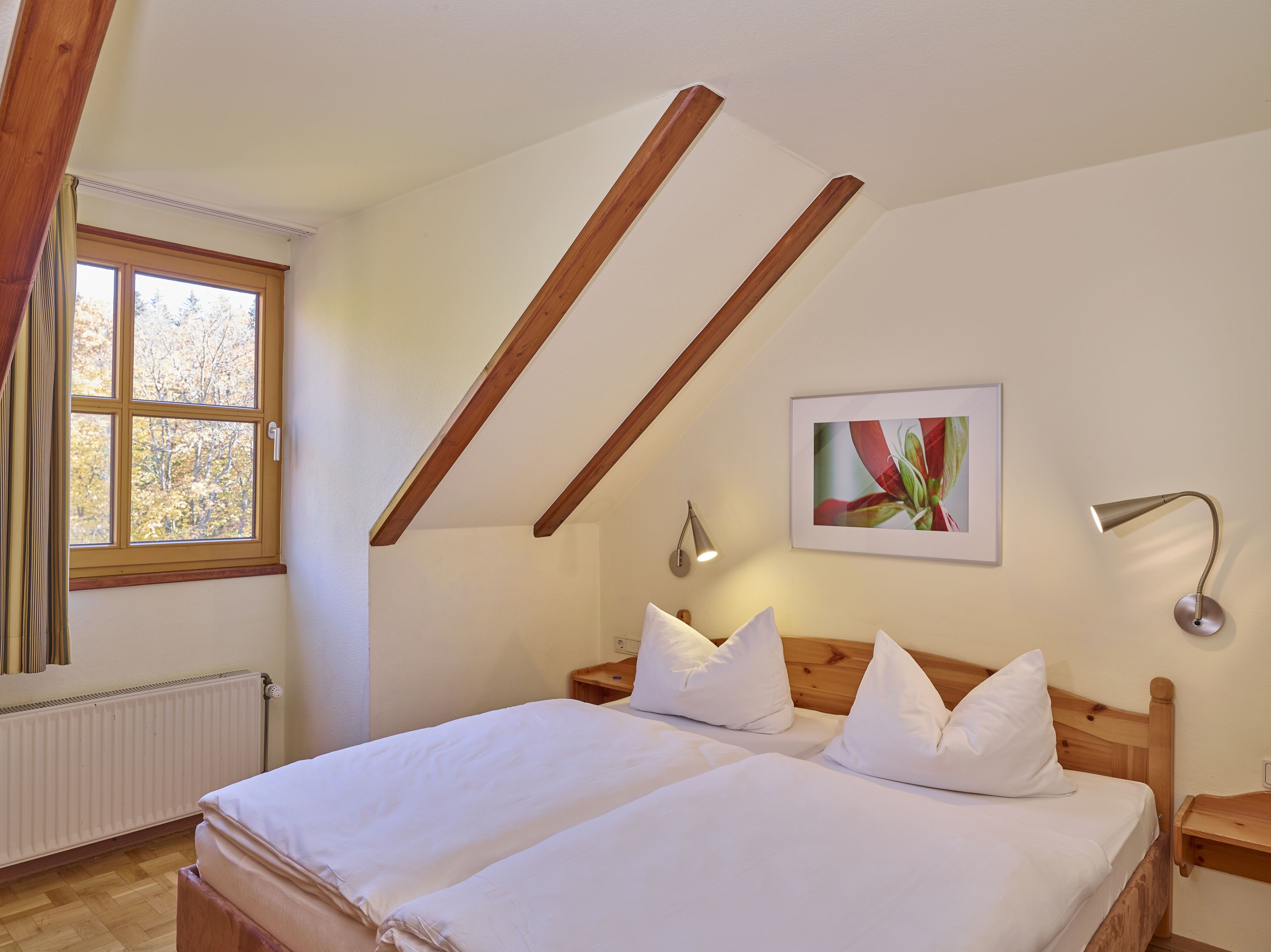 Ein Schlafzimmer mit Holzboden, einem Bett mit weißer Bettwäsche, einem Fenster mit Blick auf Bäume und einem gerahmten Bild an der Wand.