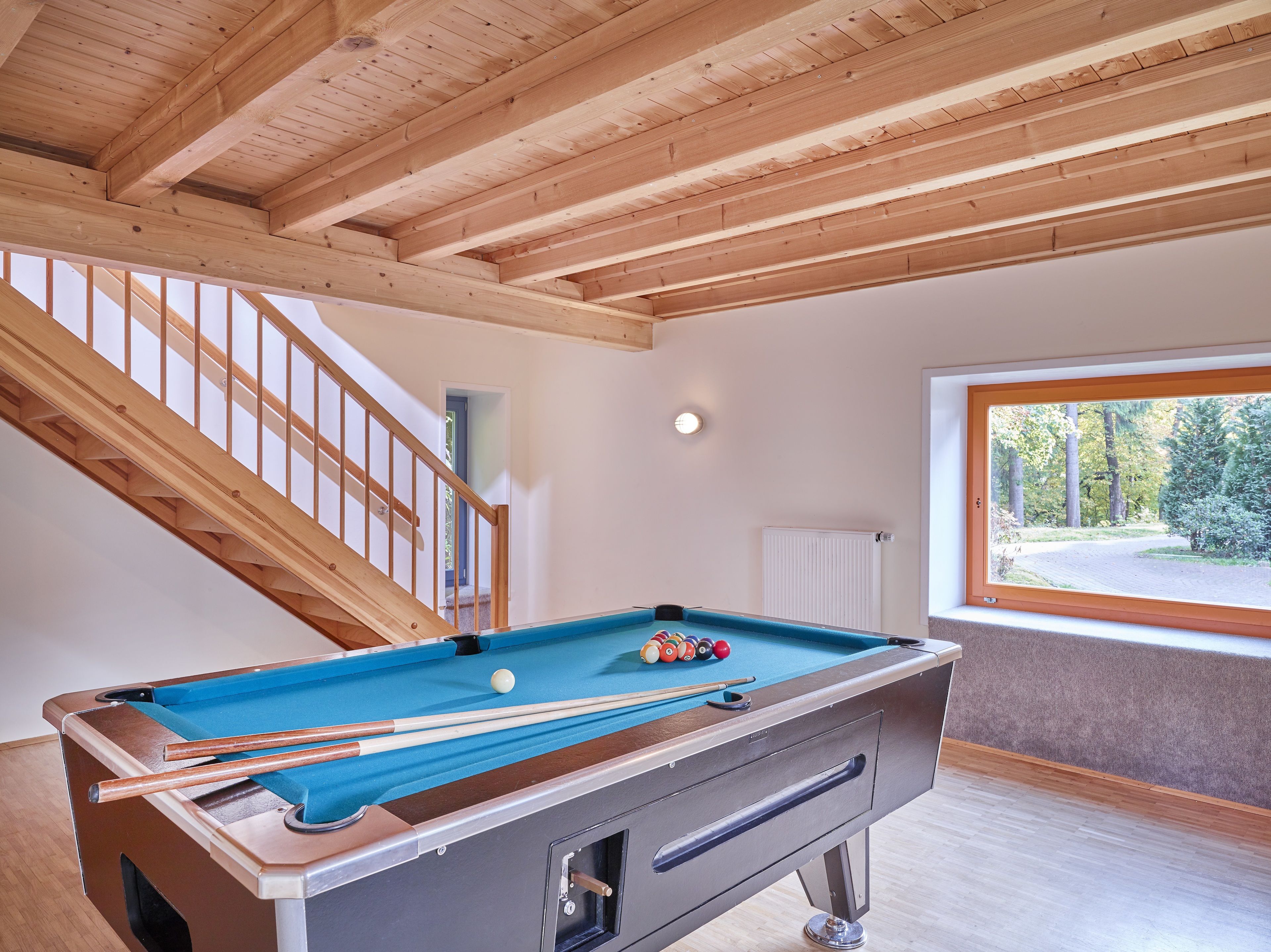 Una mesa de billar en una habitación con vigas de madera en el techo, un ventanal con vistas a árboles y una escalera.