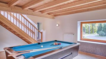 Una mesa de billar en una habitación con vigas de madera en el techo, un ventanal con vistas a árboles y una escalera.