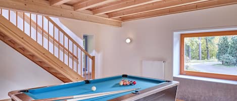 Een pooltafel in een kamer met houten balken in het plafond, een raam met uitzicht op bomen, en een trap.