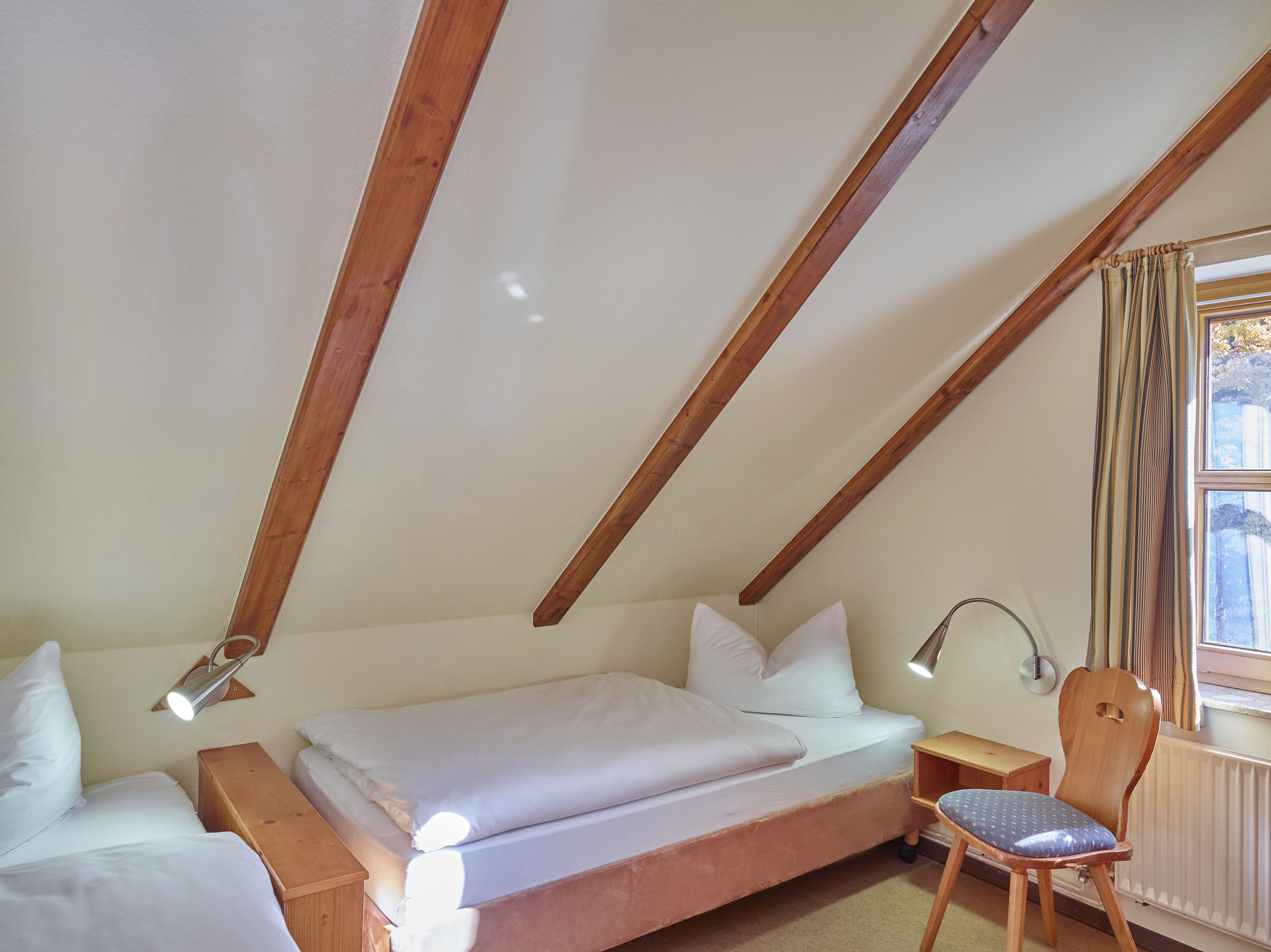 A bedroom with a sloped ceiling, wooden beams, a bed with white bedding, a bedside table with a lamp, a chair, and a window with curtains.