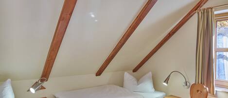Una camera da letto con soffitto spiovente, travi in legno, un letto con biancheria bianca, un comodino con una lampada, una sedia e una finestra con tende.