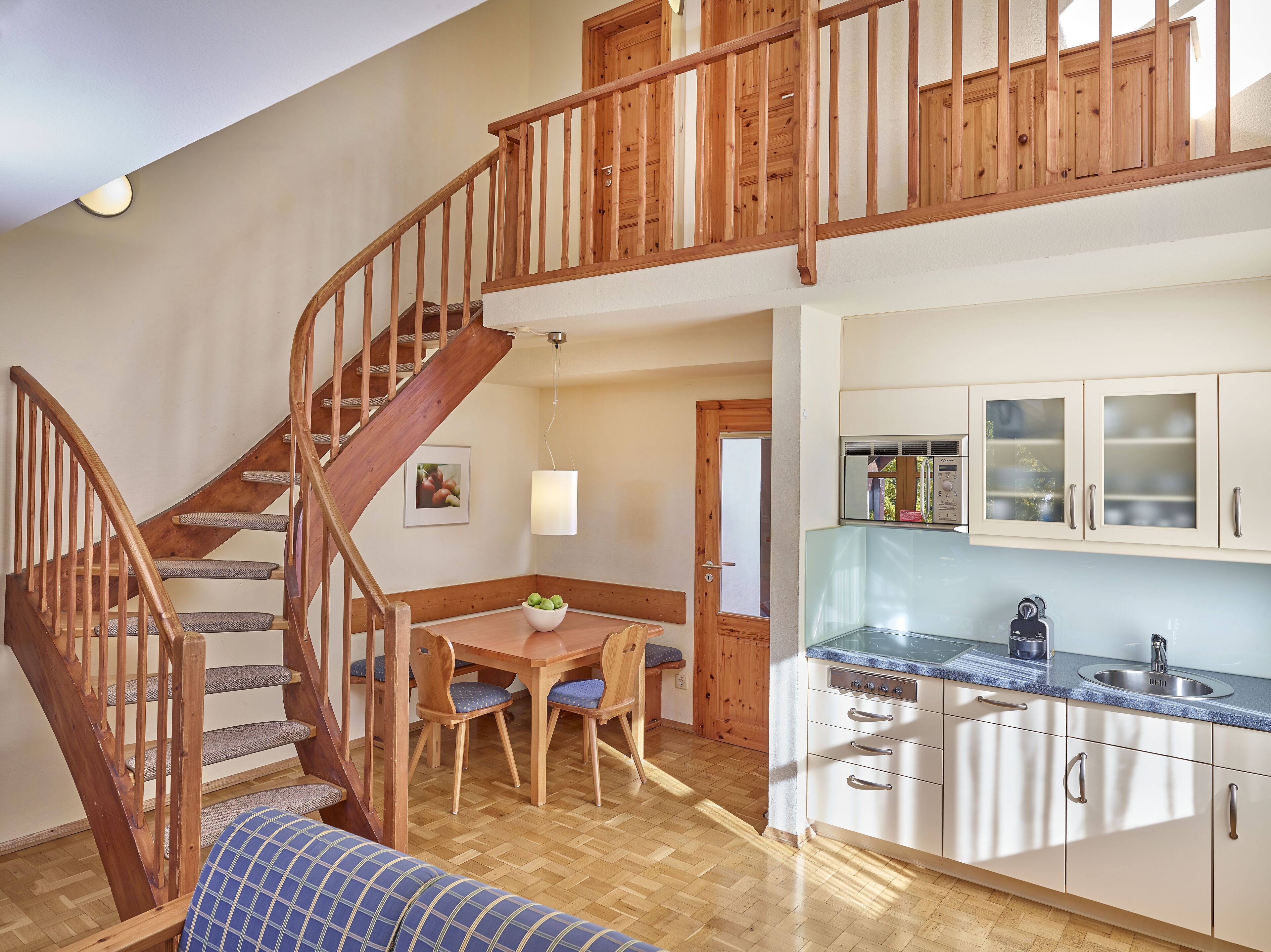 Una escalera con barandas de madera que conduce a un segundo piso, un comedor con mesa y sillas, y una cocina con armarios blancos y un lavaplatos de acero inoxidable.