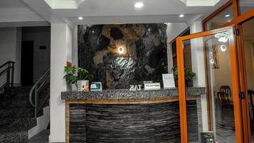 Un mostrador de recepción con un fondo de pared de piedra, una escalera a la izquierda y una puerta de cristal a la derecha.