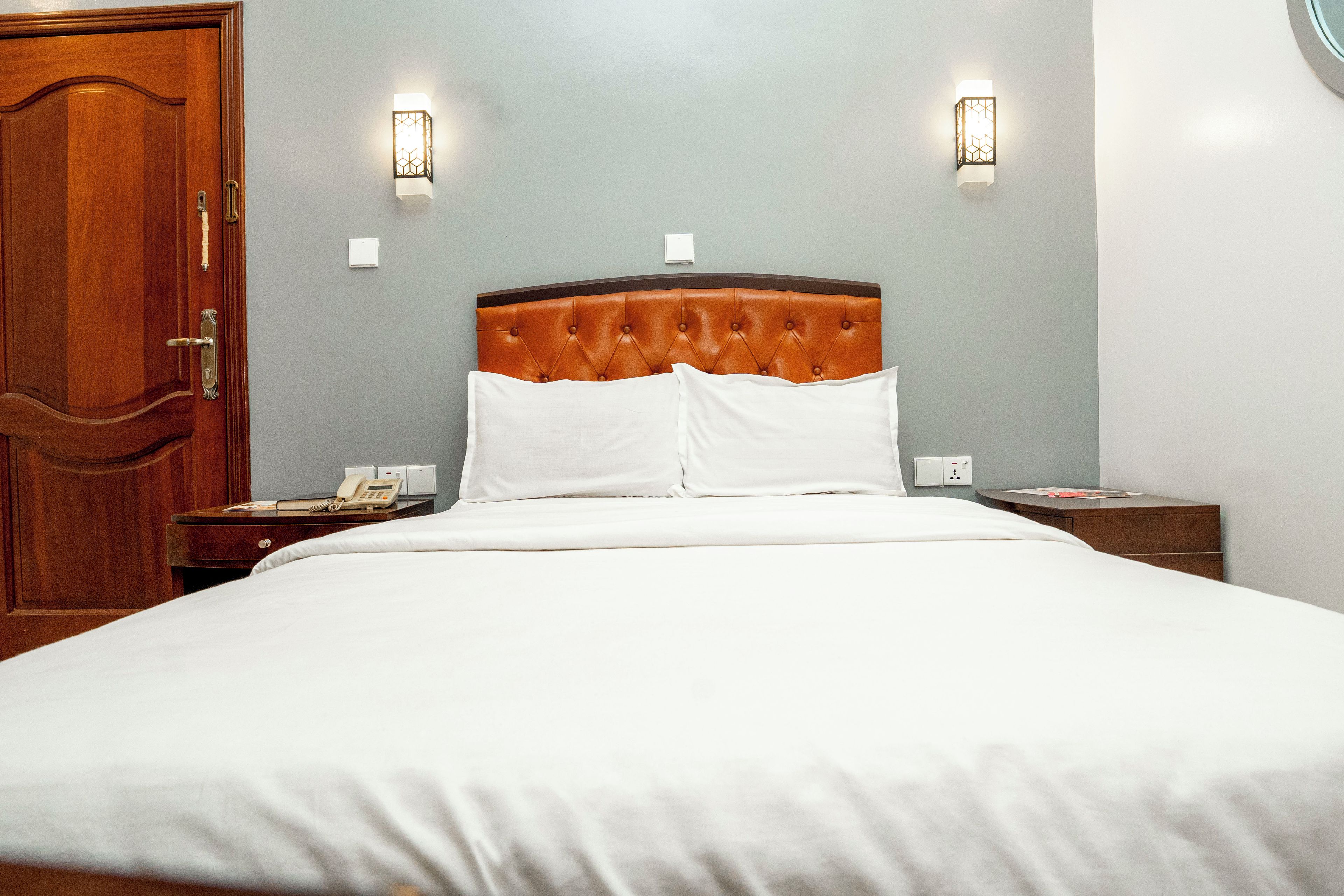 Habitación de hotel con una cama grande, una puerta de madera, dos mesitas de noche con lámparas y un teléfono sobre la mesa izquierda.