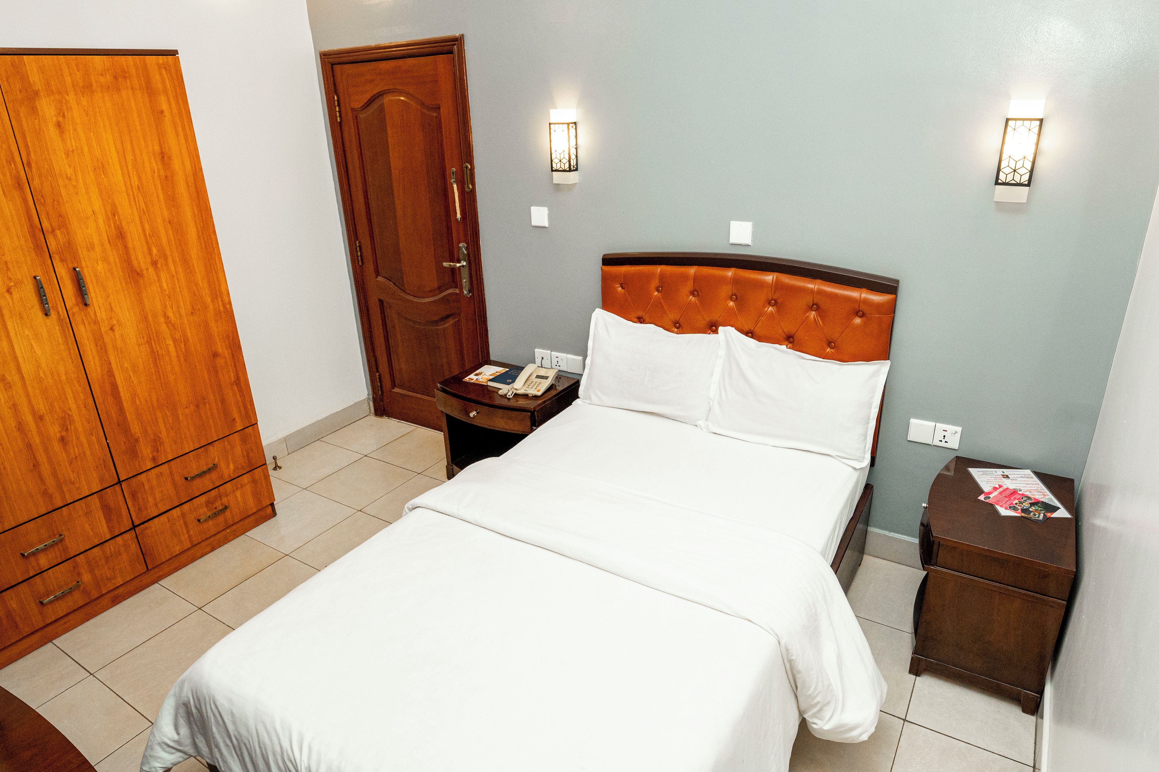 A hotel room with a bed, a wooden wardrobe, a nightstand with a telephone, and a wall-mounted light fixture.