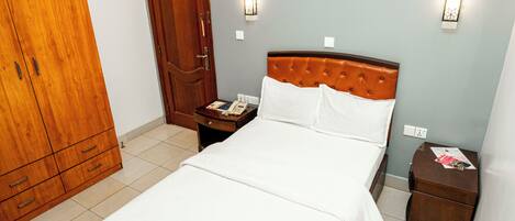A hotel room with a bed, a wooden wardrobe, a nightstand with a telephone, and a wall-mounted light fixture.