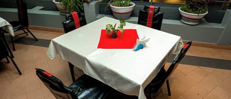 Un coin repas avec une table dressée pour deux personnes, des chaises, des plantes en pot et une serviette rouge.