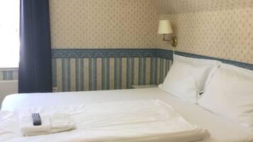 A neatly made bed with white linens, a remote control, and a folded towel on a hotel room with patterned wallpaper and a blue-striped border.