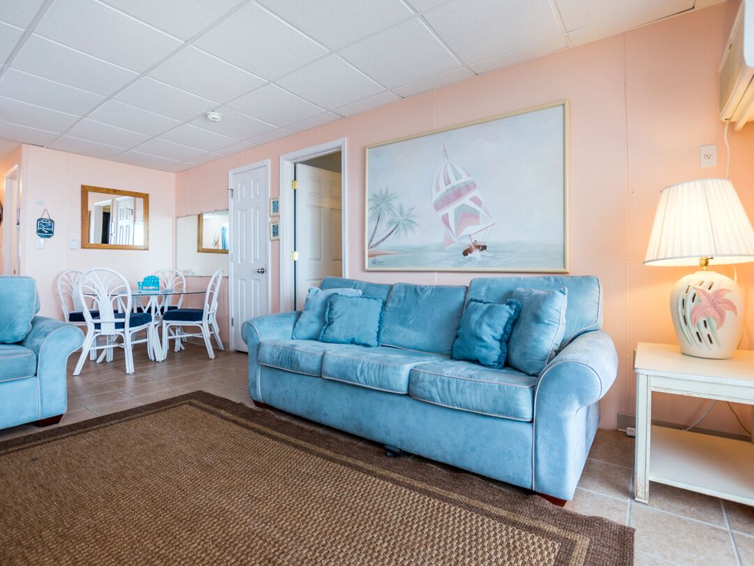Ein Wohnzimmer mit einem hellblauen Sofa, einem Esstisch mit Stühlen, einem Segelbootgemälde und einer Lampe auf einem Beistelltisch.