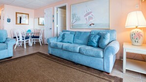 A living room with a light blue sofa, a dining table with chairs, a painting of a sailboat, and a lamp on a side table. - Oceanfront condos with rare 2 night minimum rentals and linens included! (Ocean City)