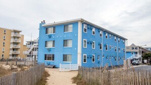 A multi-story building with a blue facade, a sandy pathway leading to the entrance, and a white fence surrounding the property. - Oceanfront condos with rare 2 night minimum rentals and linens included! (Ocean City)
