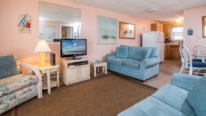 A living room with a television, a sofa, a small table with a lamp, and a kitchen area in the background. - Oceanfront condos with rare 2 night minimum rentals and linens included! (Ocean City)