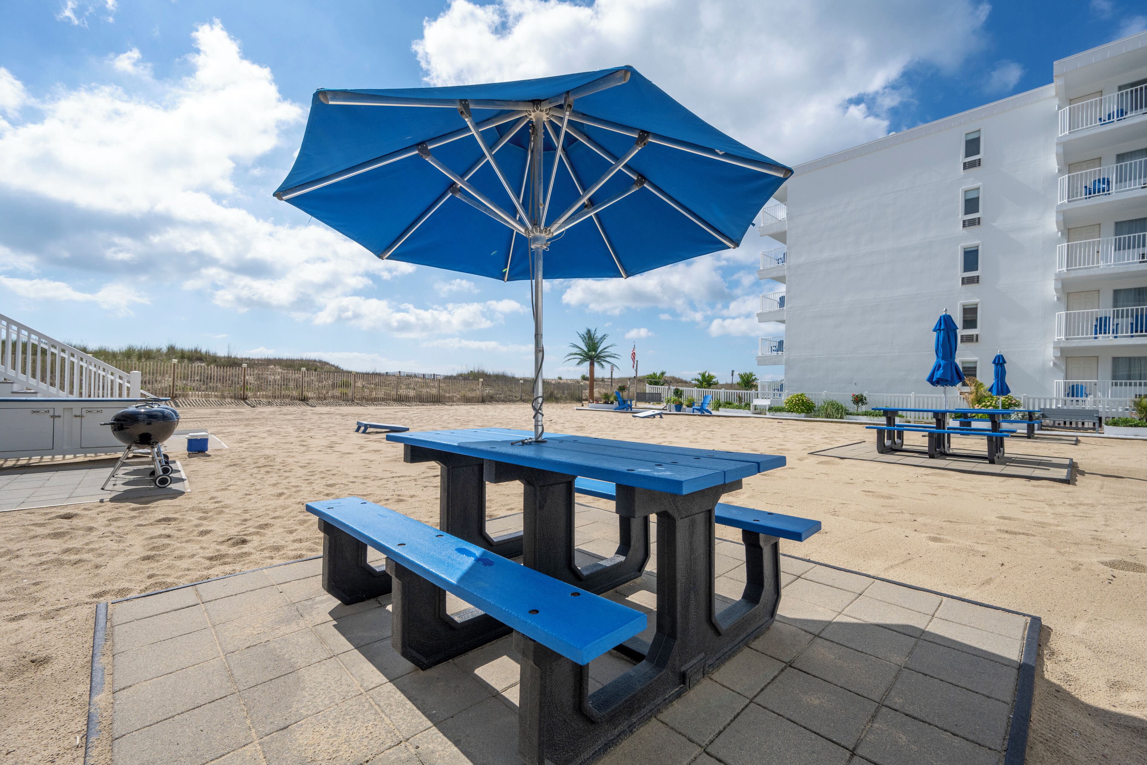 Un patio frente a la playa con una sombrilla azul, una mesa de picnic y una parrilla.