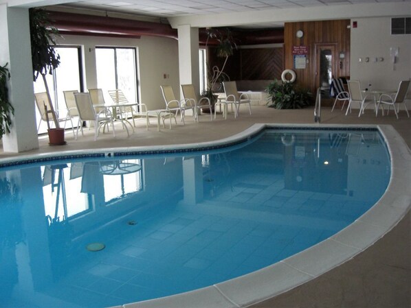 An indoor swimming pool with clear blue water, surrounded by white lounge chairs and a few potted plants.